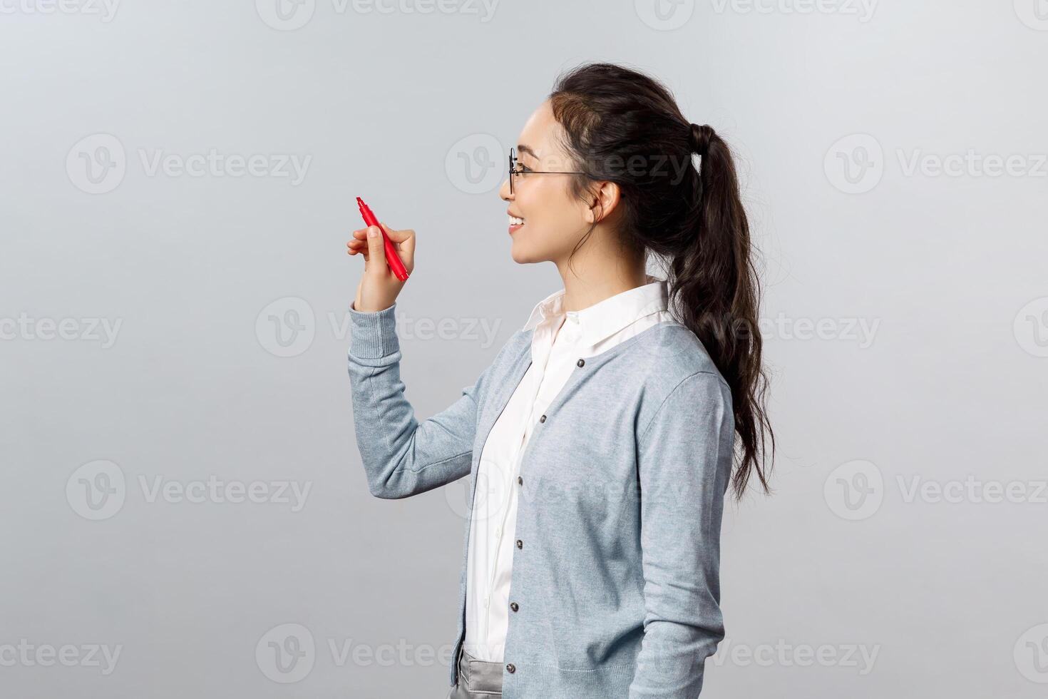 Teaching, online education and lifestyle concept. Smart young female teacher writing down on blackboard, explain exercise to students studying on distance, smiling as holding pen and look left photo