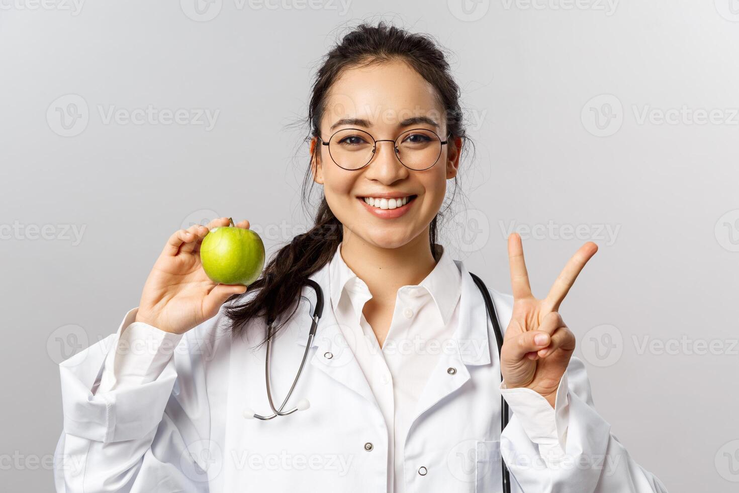 Portrait of enthusiastic young happy smiling female doctor in white coat, show peace sign and green apple, asking people stay healthy, eat more fruits and vitamins, grey background photo