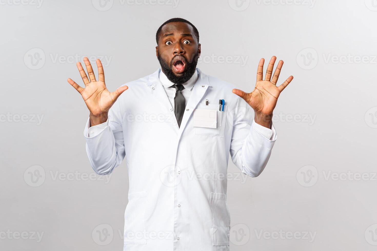 Portrait of shocked and nervous african-american male doctor raising hands in surrender, trying calm down tensed family of patient, look insecure and unaware what do, stand grey background photo