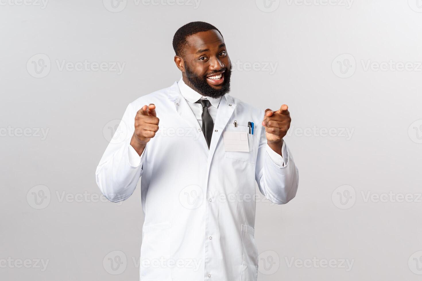 Portrait of friendly handsome doctor pointing at camera, inviting people for check-up, telling stay home be safe during corona, covid19 pandemic, smiling encourage vaccinate, grey background photo