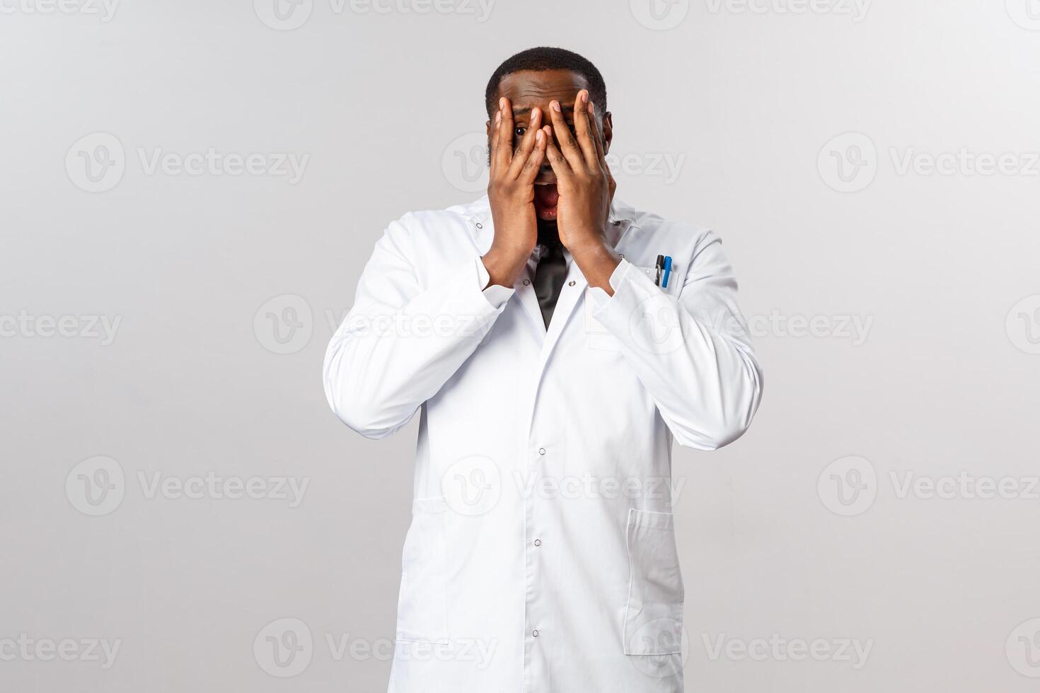 Scared, insecure and shocked african-american male doctor, physician cant look at severe trauma of patient, close eyes and peeking through fingers with frightened, horrified expression photo