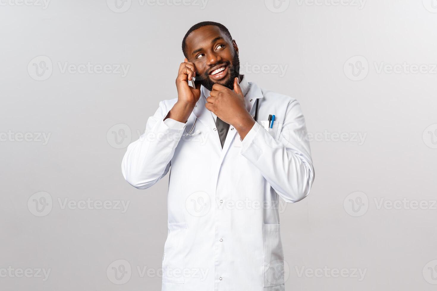 Covid19, pandemic, online medicine concept. Portrait of handsome happy, smiling doctor, male phycisian touch beard thoughtful, talking on phone, consult patient about treatment on social-distancing photo