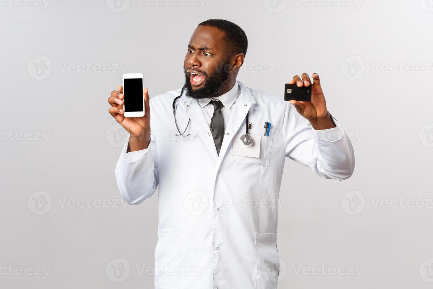 Covid19, pandemic and online medicine concept. Complaining angry african-american man looking at mobile phone display, hold credit card, outraged with huge bills and expensive app subscribtion photo
