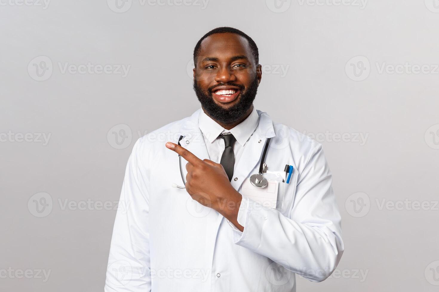 Covid19, hospital check-up and healthcare concept. Handsome enthusiastic african-american bearded doctor explain how to follow prescribtion at hospital, invite patient to clinic, point left photo