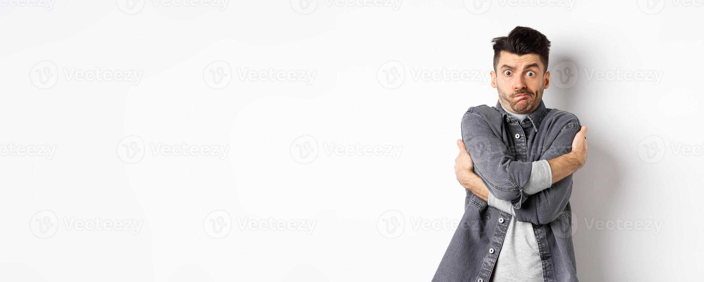 Funny young man hugging himself and grimacing, feeling embarrassed and exposed, covering body from people glances, white background photo