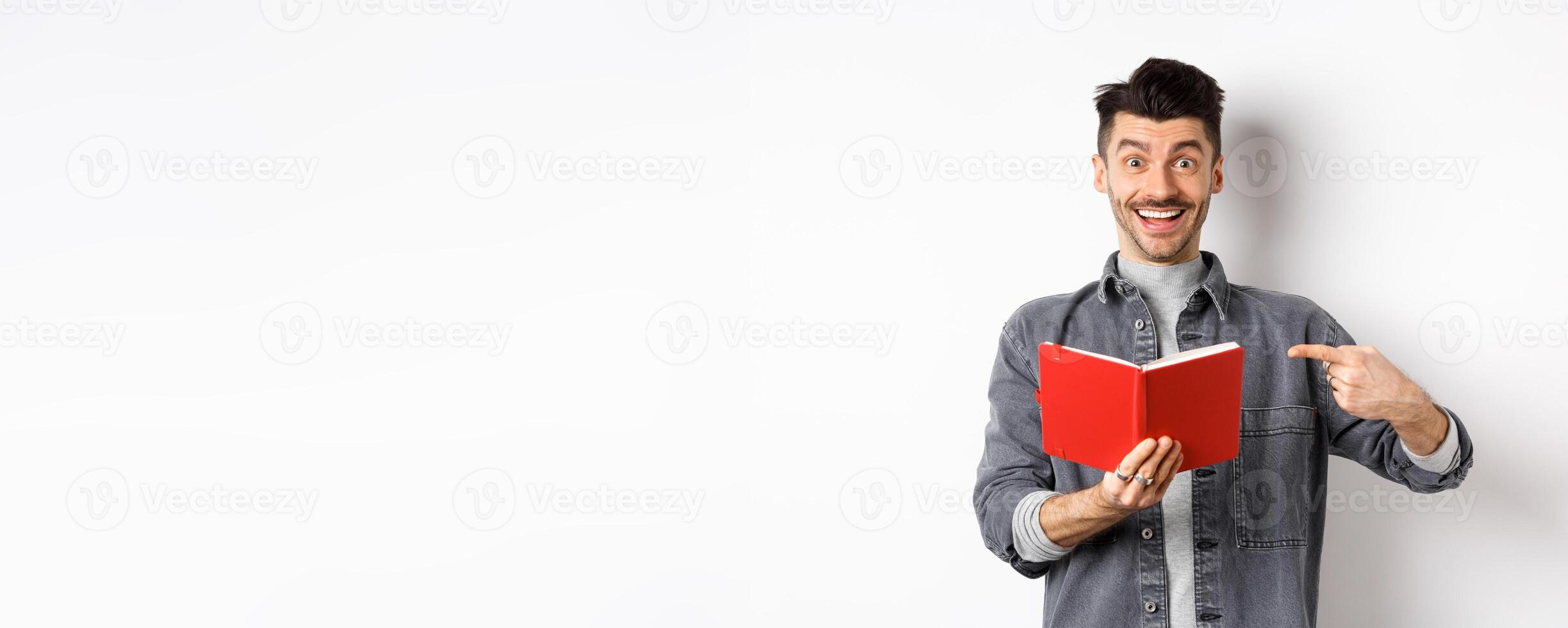 Happy smiling guy pointing at red planner, showing good journal or book, standing on white background photo
