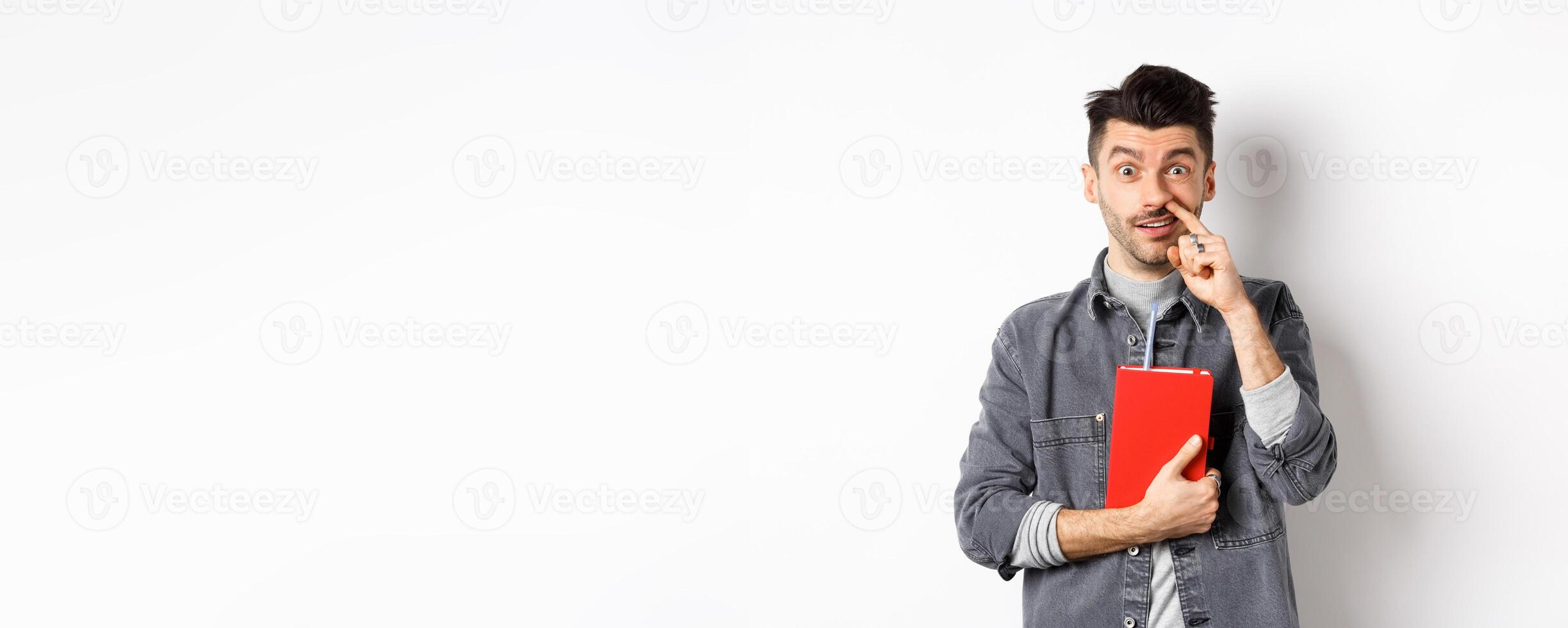 Young guy picking nose and holding book, look stupid at camera, standing against white background photo