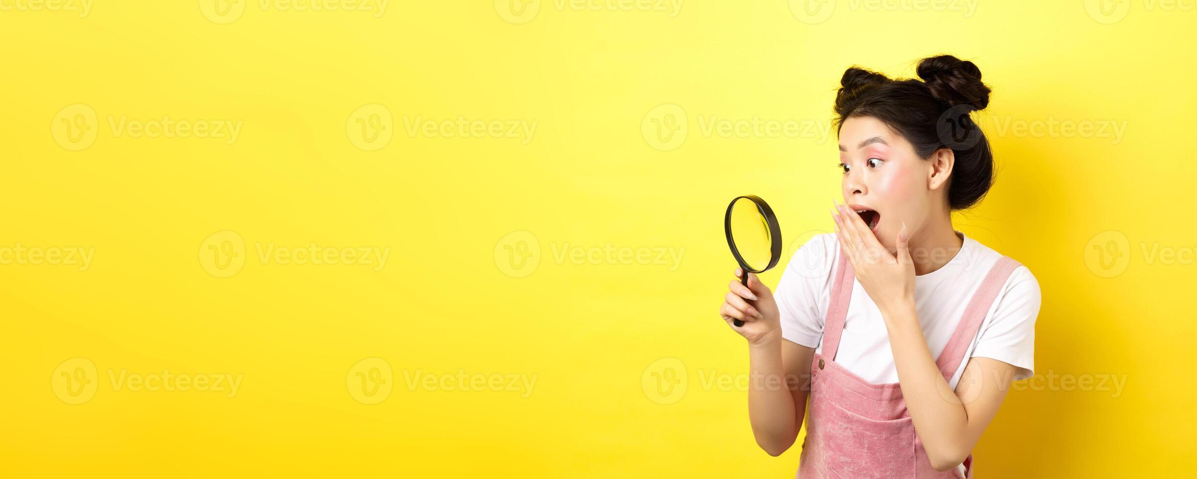 Excited silly asian girl gasping wondered, found something interesting, looking through magnifying glass, standing on yellow background photo