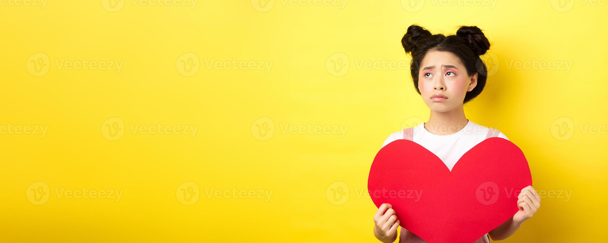 Sad and lonely asian girl standing in make-up with big red heart cutout, left alone on Valentines day, looking left disappointed or upset, standing on yellow background photo
