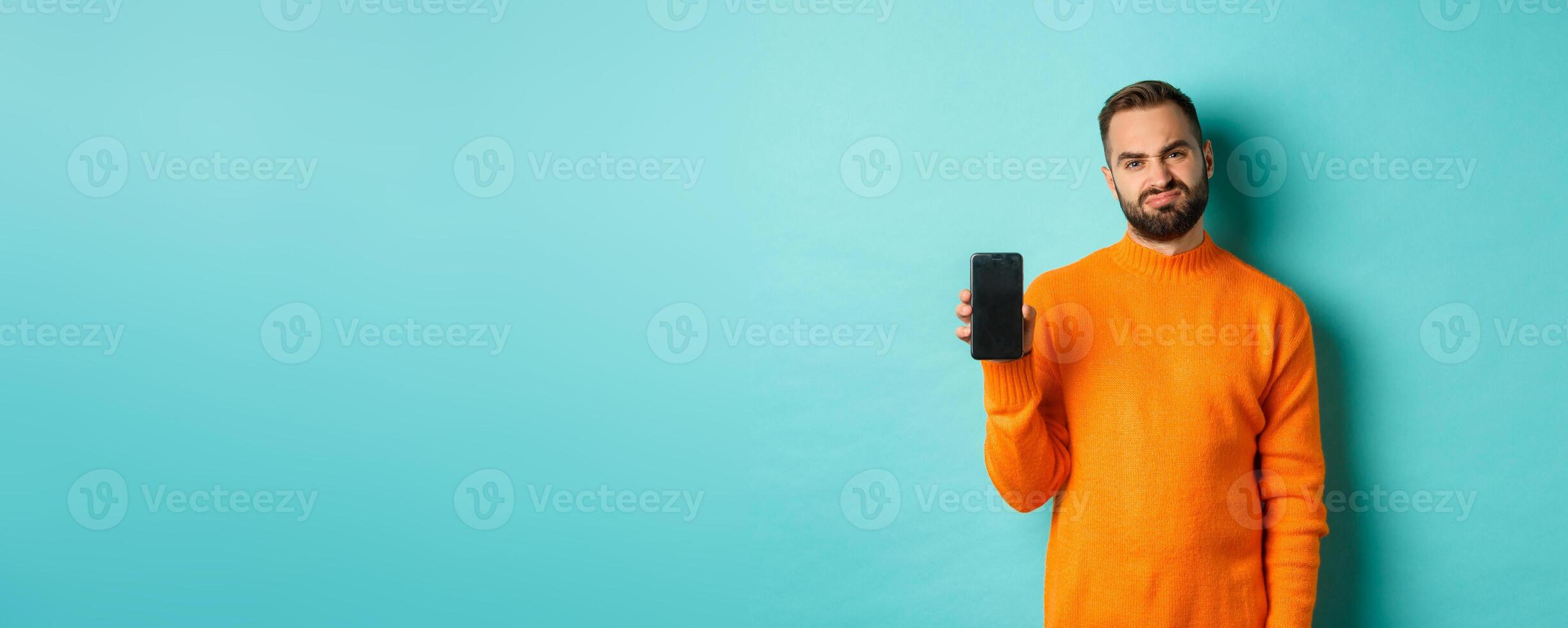 Displeased and disappointed man in orange sweater, grimacing and showing mobile screen, standing over light blue background photo