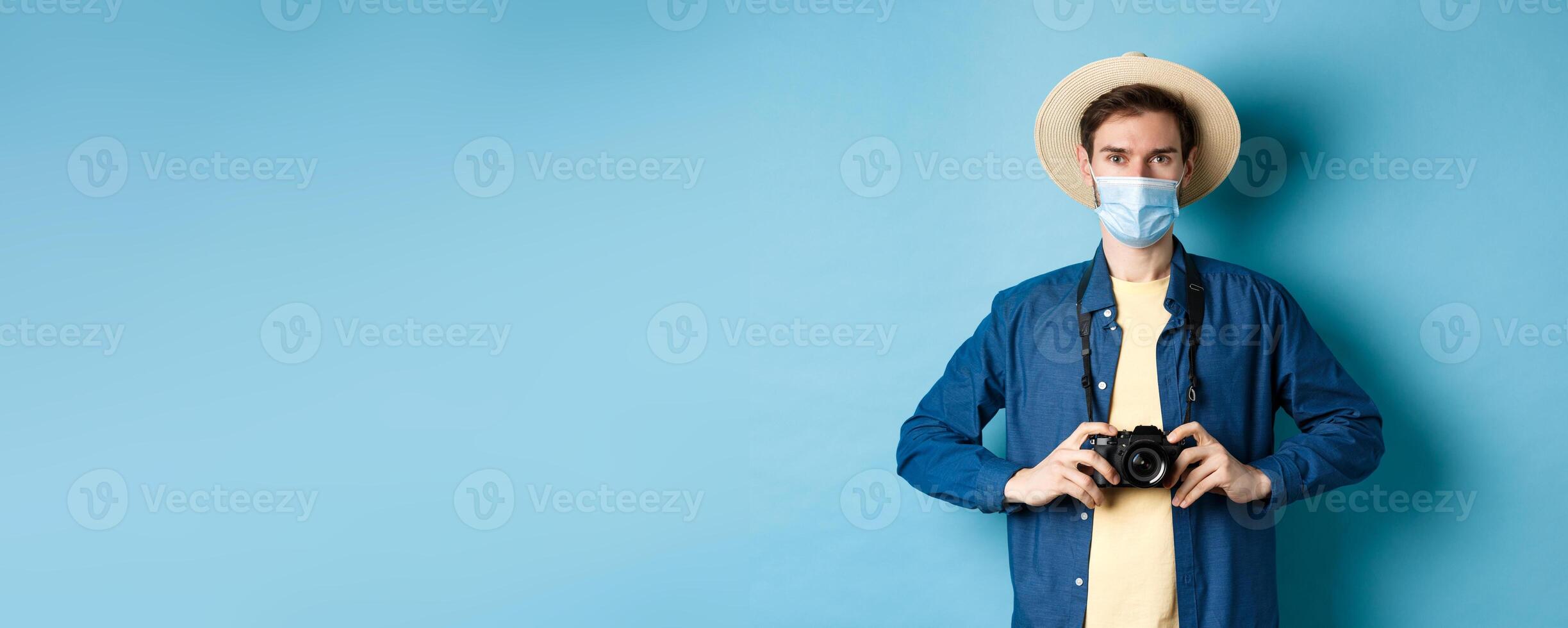 Covid-19, pandemic and travel concept. Young guy travelling abroad with camera, taking pictures on vacation, wearing medical mask from coronavirus, blue background photo