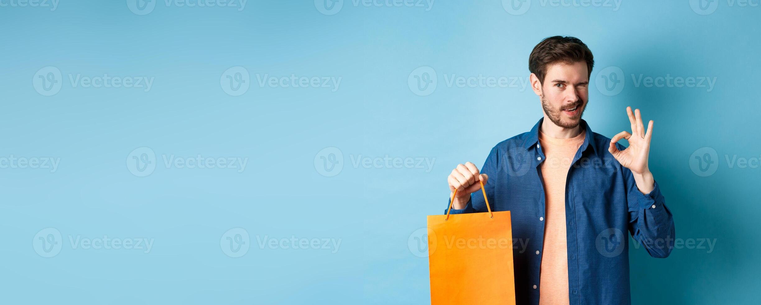 Modern man in casual outfit showing shopping bag and okay sign, winking at camera, recommending shop, standing on blue background photo