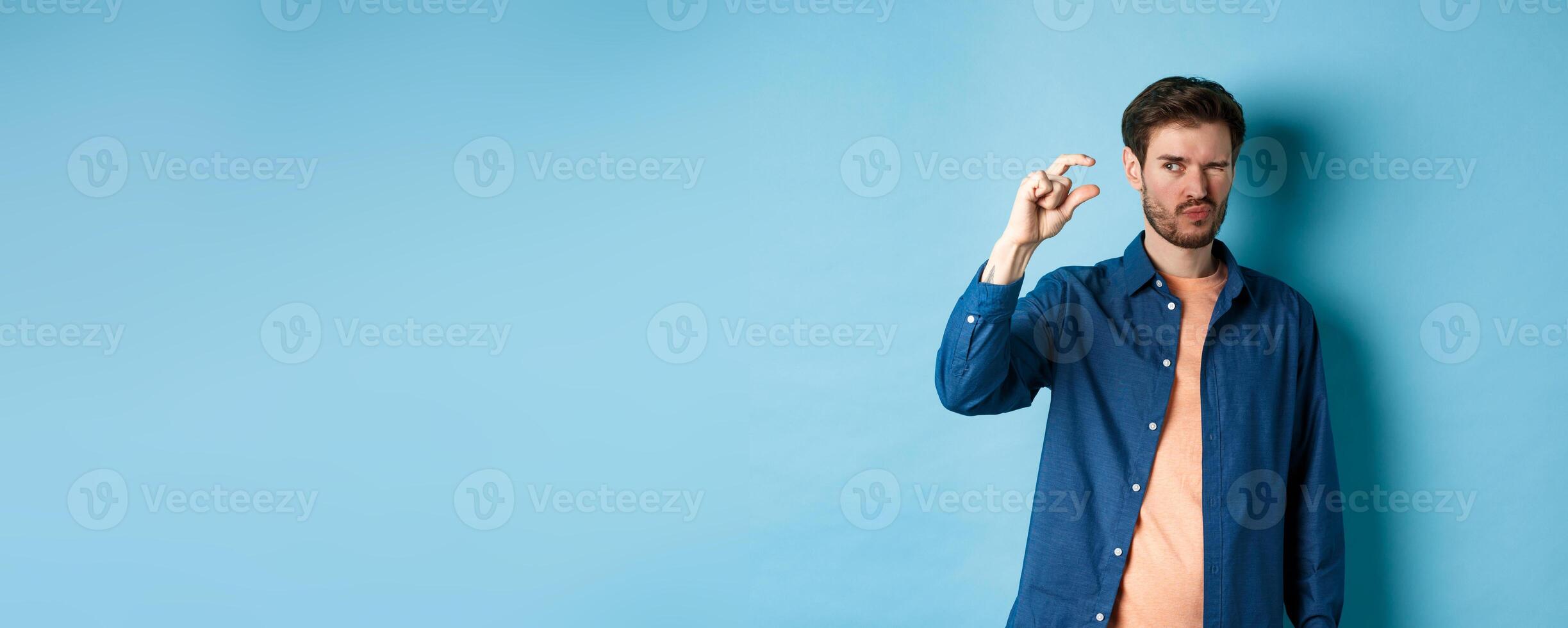 Image of young caucasian man showing small size and squinting to see something tiny, standing on blue background photo