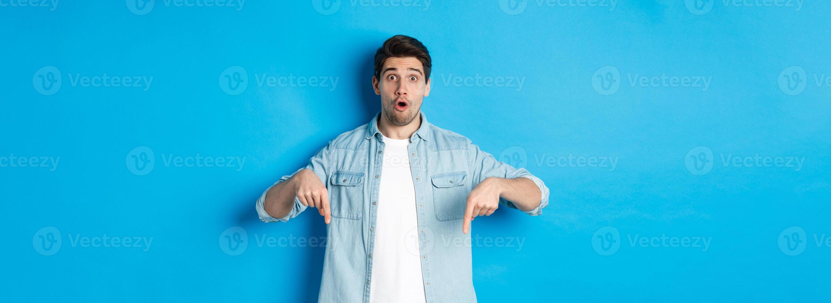 Surprised bearded adult man in casual outfit, pointing fingers down and looking startled, asking question about promotion, standing over blue background photo