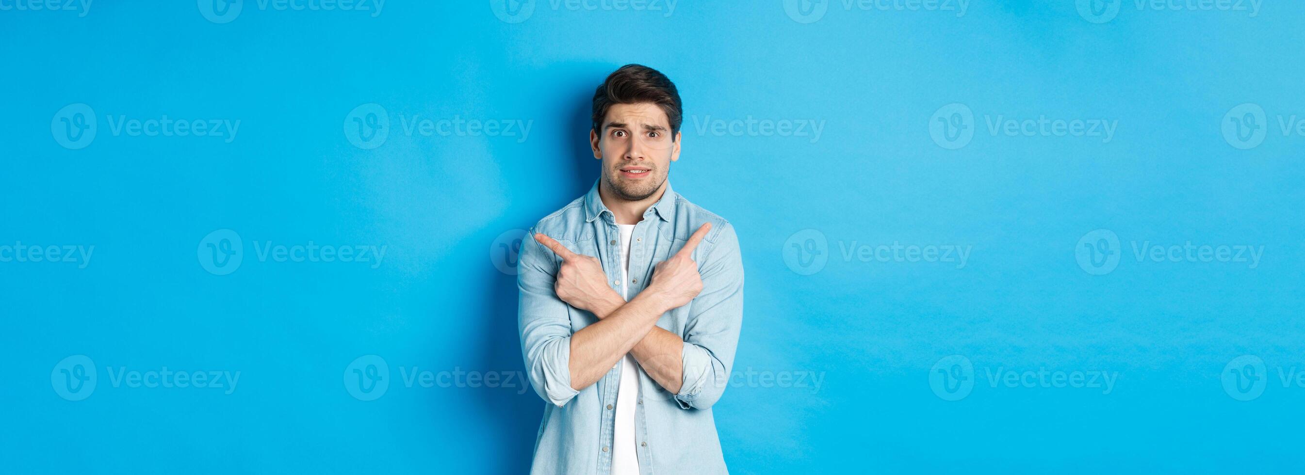 Portrait of nervous guy pointing fingers sideways, looking indecisive and asking for help with choice, showing left and right promo offers, standing against blue background photo