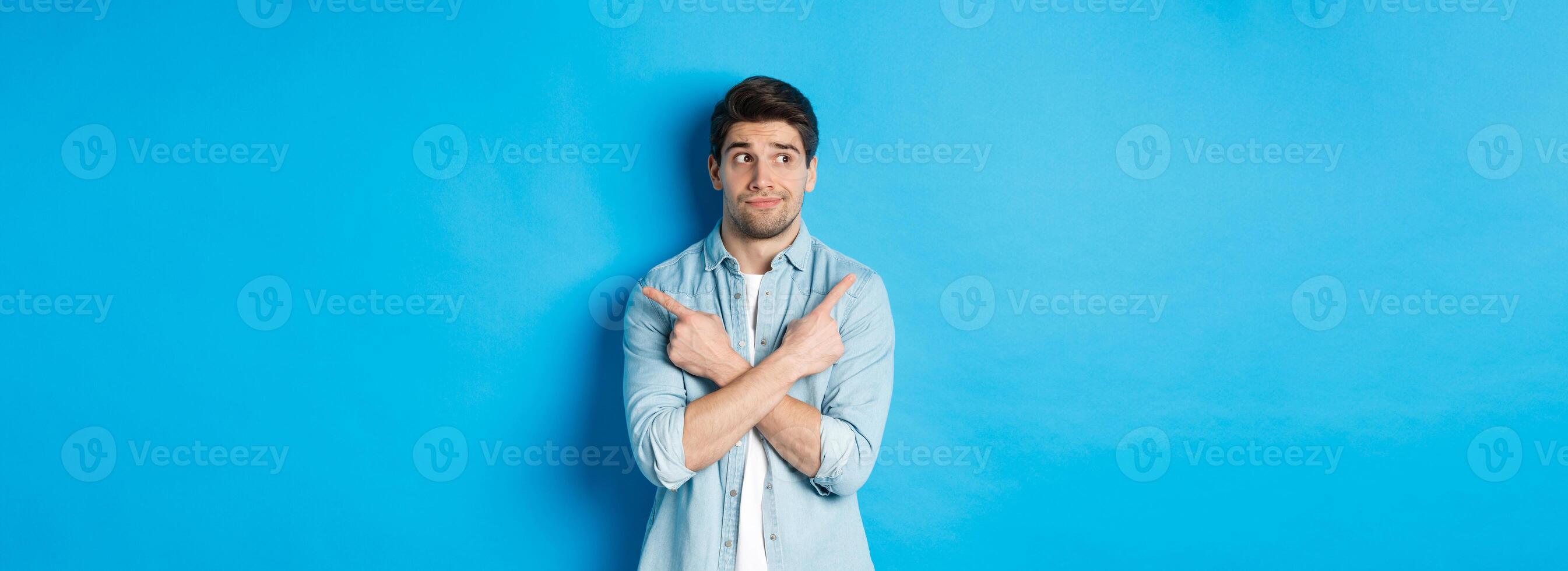 Portrait of indecisive adult man pointing fingers sideways but looking left, making choice between two products, standing against blue background photo
