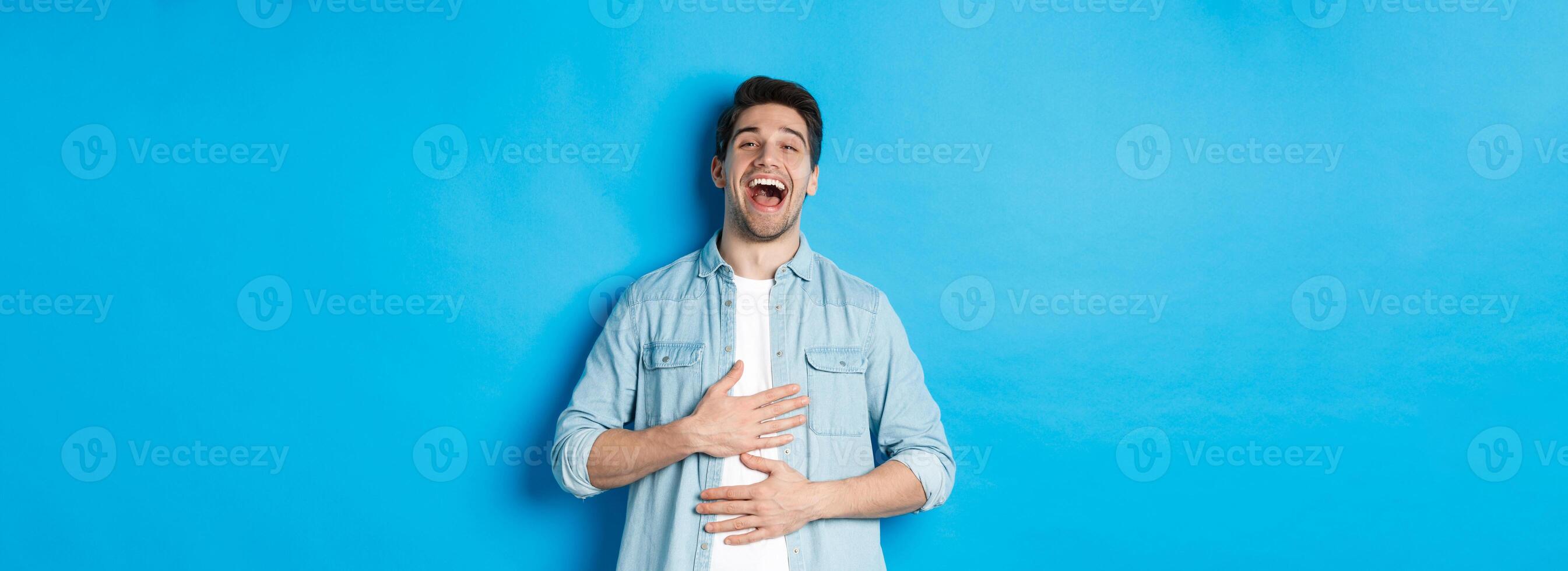 Happy handsome 30 year old man touching belly, laughing out loud and looking at something funny, standing against blue background photo