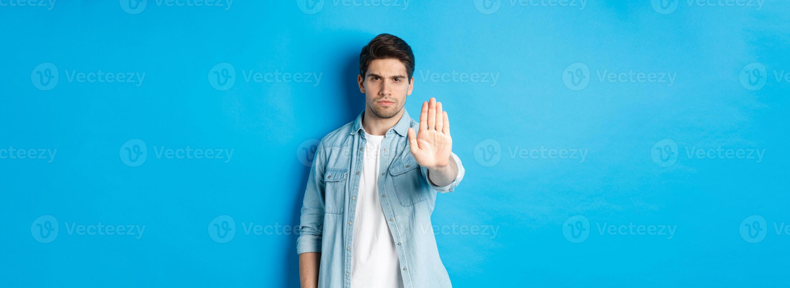 Serious man frowning and saying no, extending hand to shop stop sign, prohibit action, standing against blue background photo