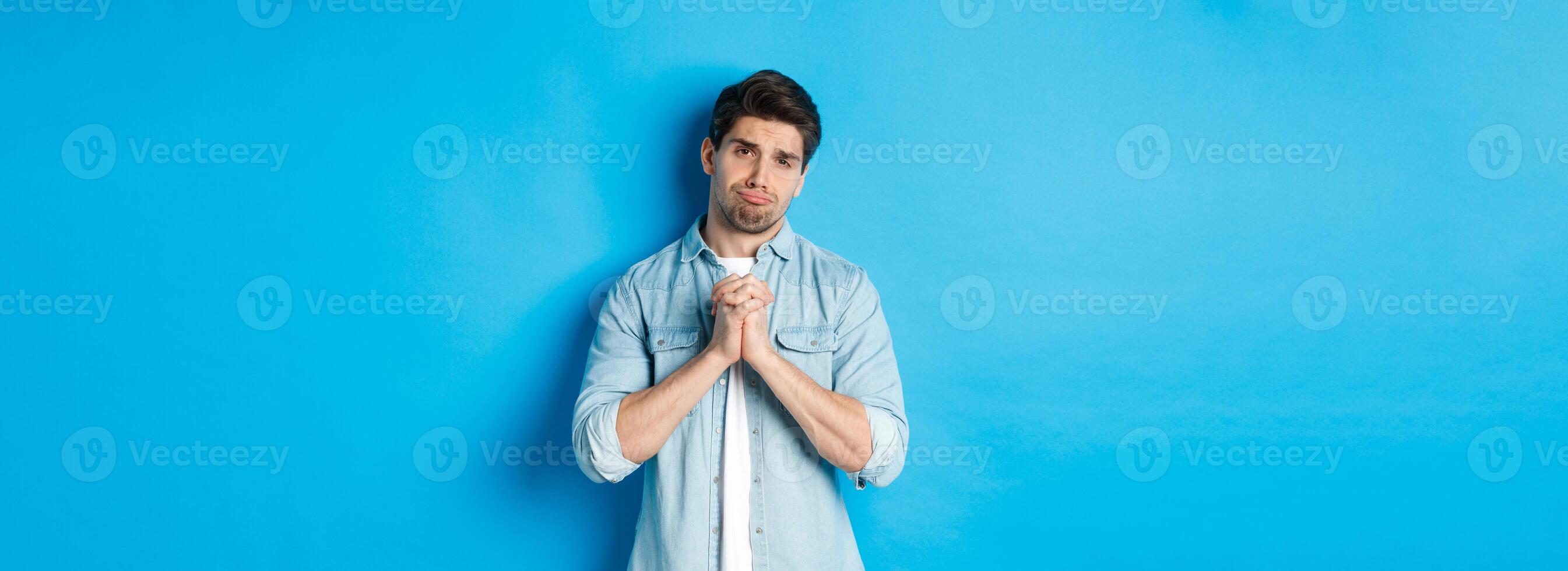 Young guy begging for help, pouting and looking sad while asking for favour, need something, standing against blue background photo