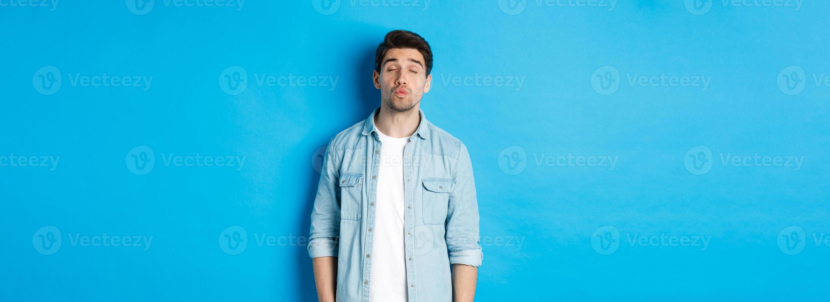 Handsome man waiting for kiss, pucker lips and close eyes while standing against blue background photo
