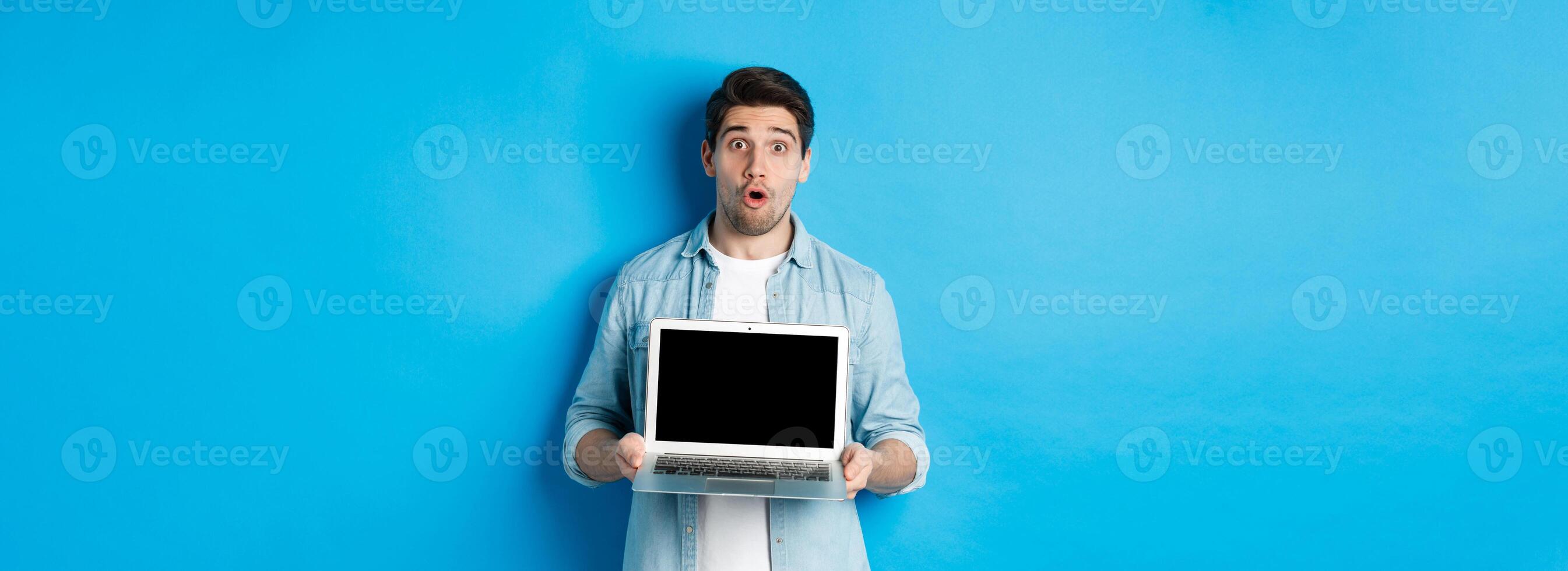 Man showing advertisement on laptop screen and looking amazed, saying wow and looking at camera, standing against blue background photo