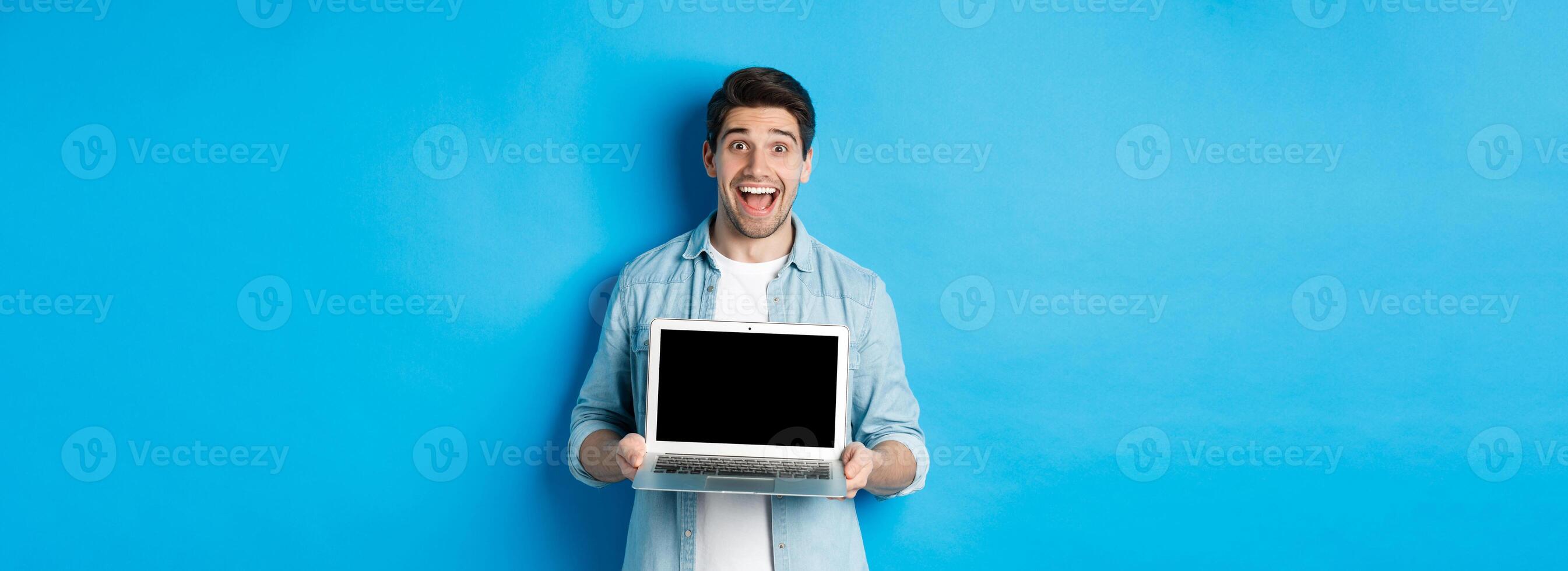 Cheerful smiling man making presentation, showing laptop screen and looking happy, standing over blue background photo