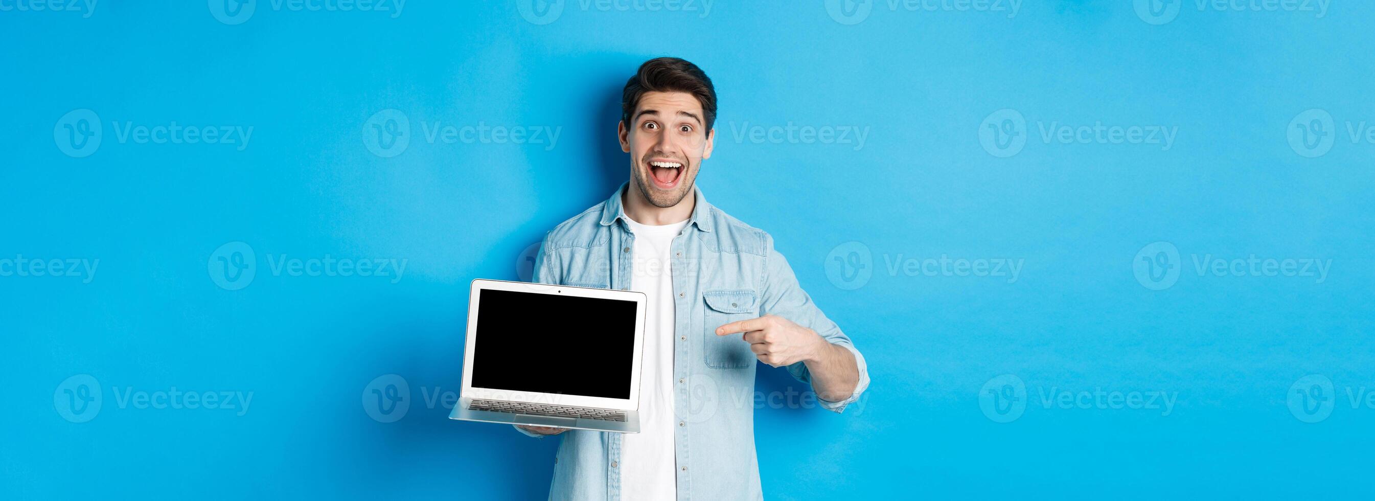Excited smiling man pointing at laptop screen, showing internet promo offer or website advertisement, standing over blue background photo