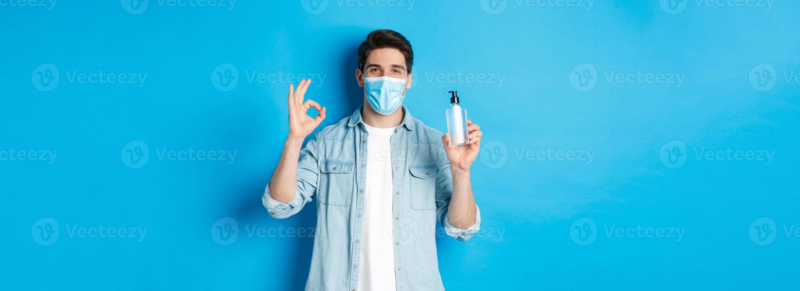 Concept of covid-19, pandemic and social distancing. Satisfied young man in medical mask recommending hand sanitizer, showing ok sign and antiseptic, standing against blue background photo