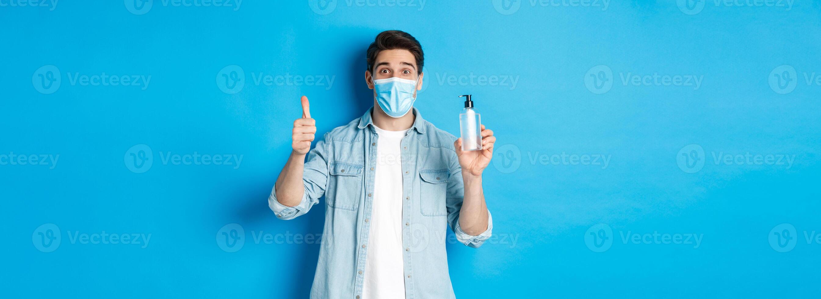 Concept of covid-19, pandemic and social distancing. Cheerful man in medical mask, showing hand sanitizer and thumbs-up, recommending sanitizer, standing against blue background photo