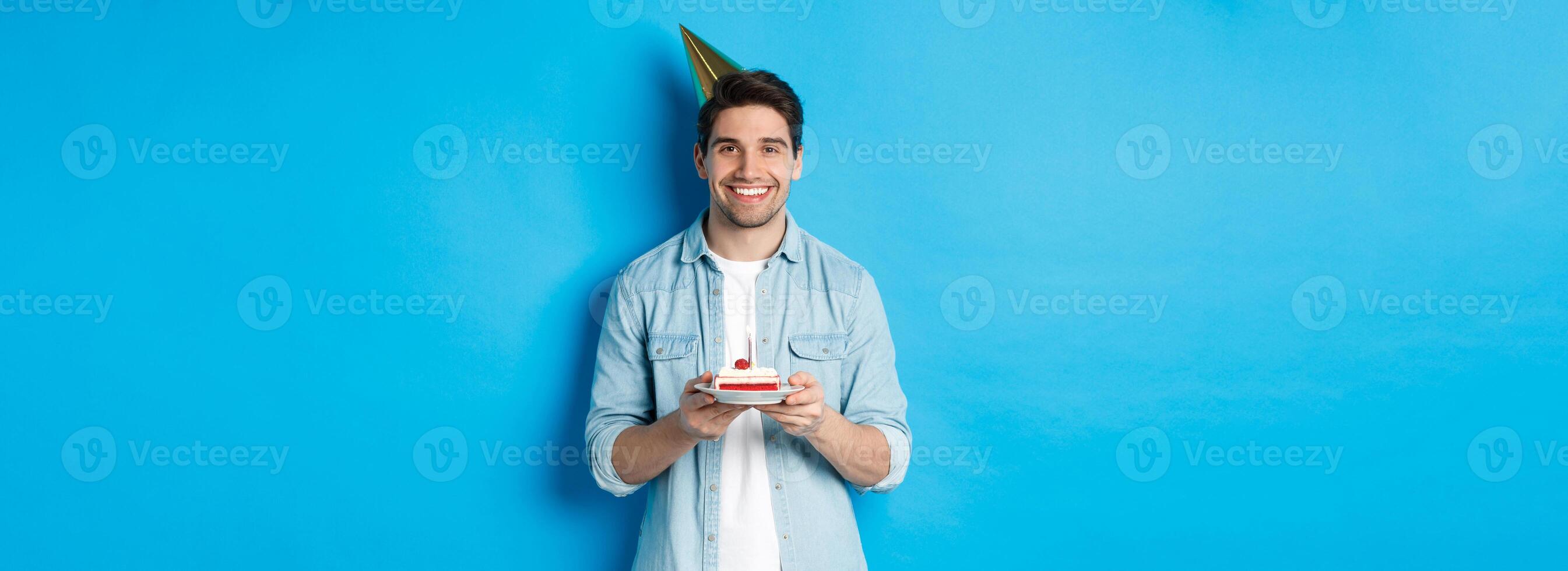 Smiling man holding b-day cake and wearing birthday party hat, celebrating over blue background photo