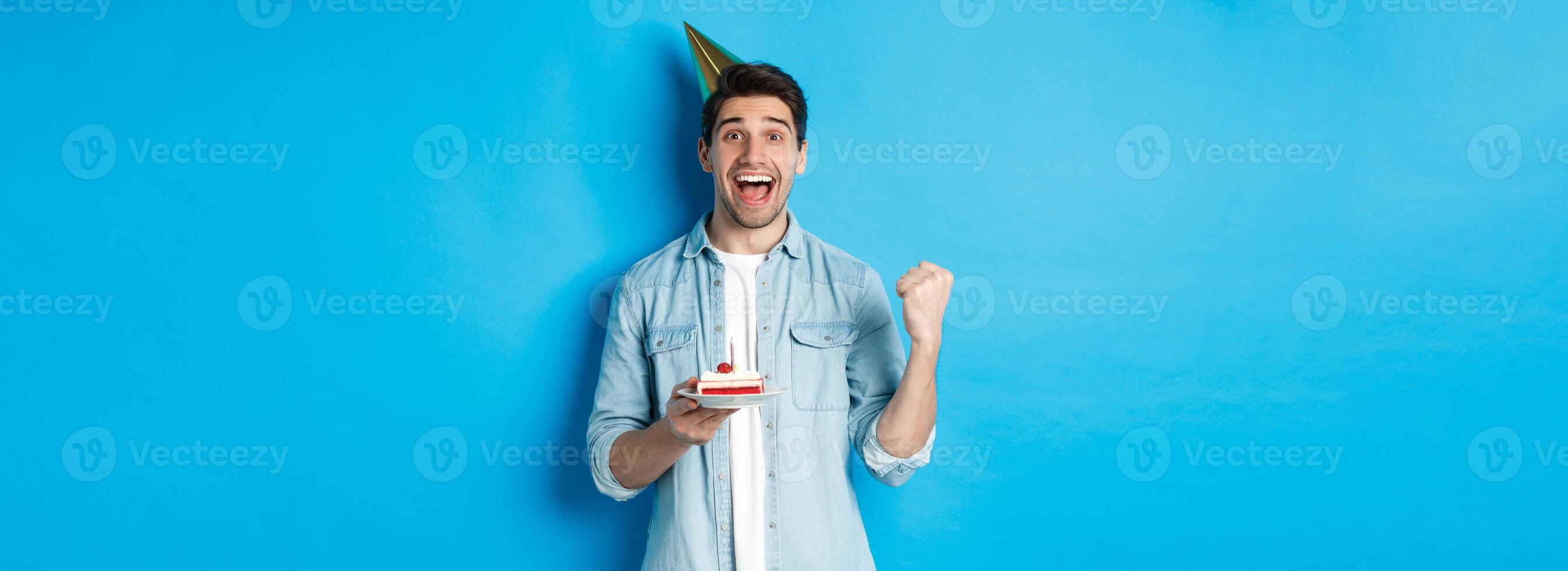 Cheerful man looking happy, celebrating birthday in party hat, holding b-day cake and make fist pump, standing over blue background photo