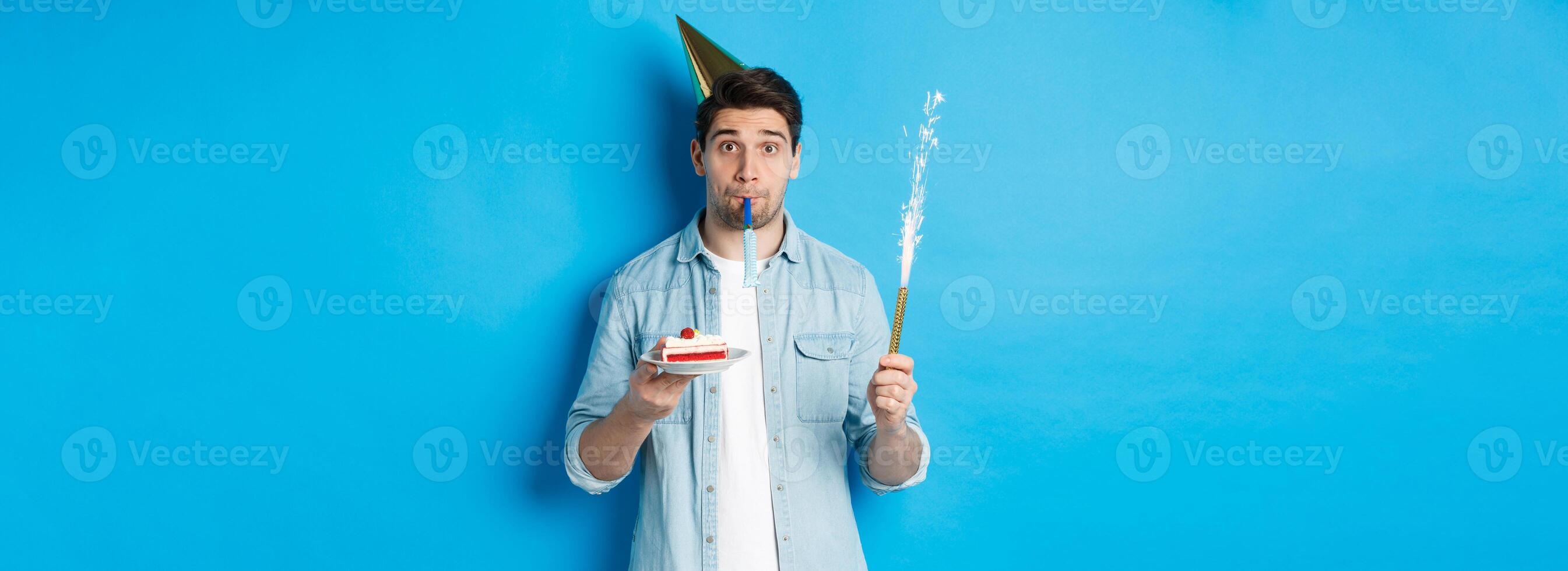 Funny guy celebrating birthday, holding b-day cake, firework and wearing party hat, standing over blue background photo