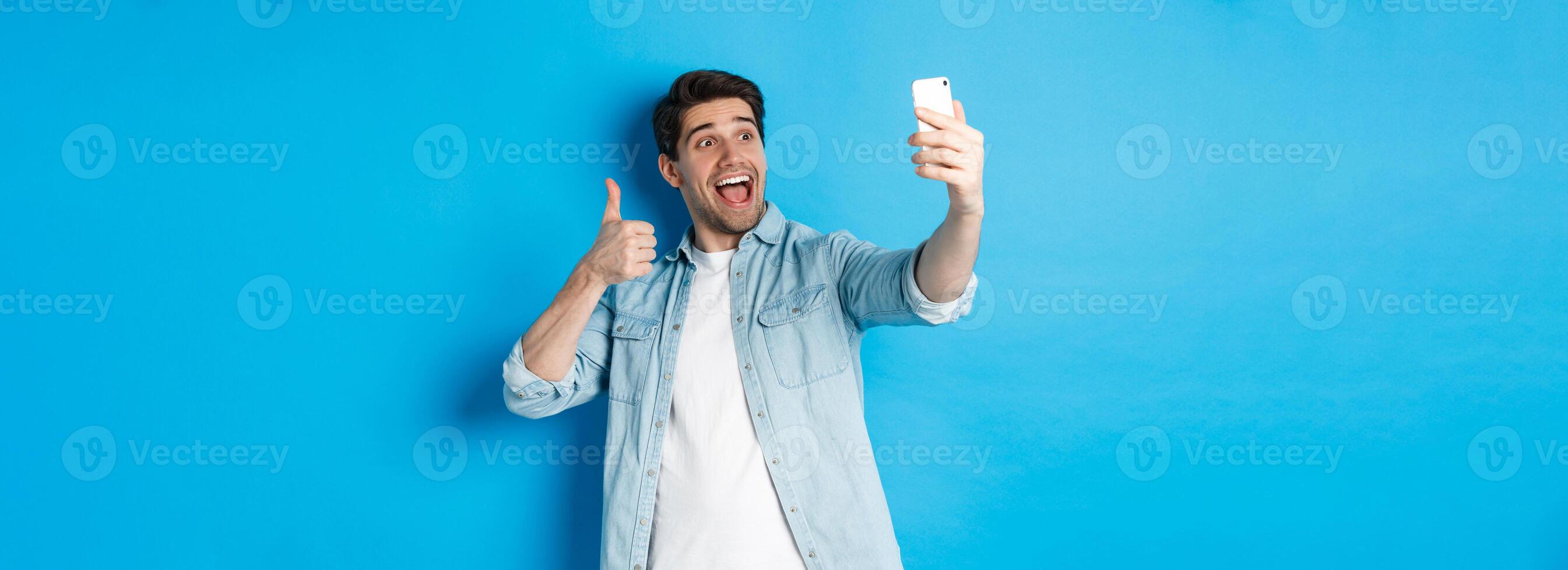 Happy man taking selfie and showing thumb up in approval on blue background, holding mobile phone photo