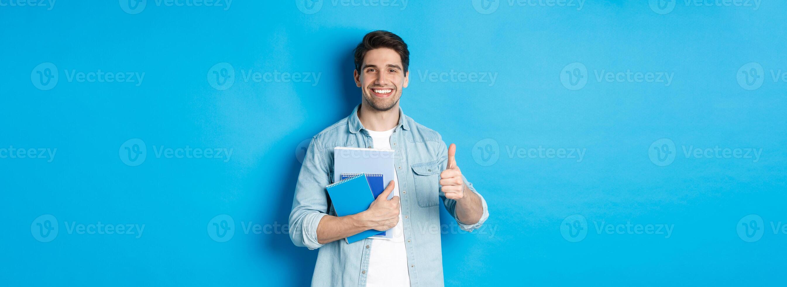 Young man student with notebooks, showing thumb up in approval, smiling satisfied, blue studio background photo