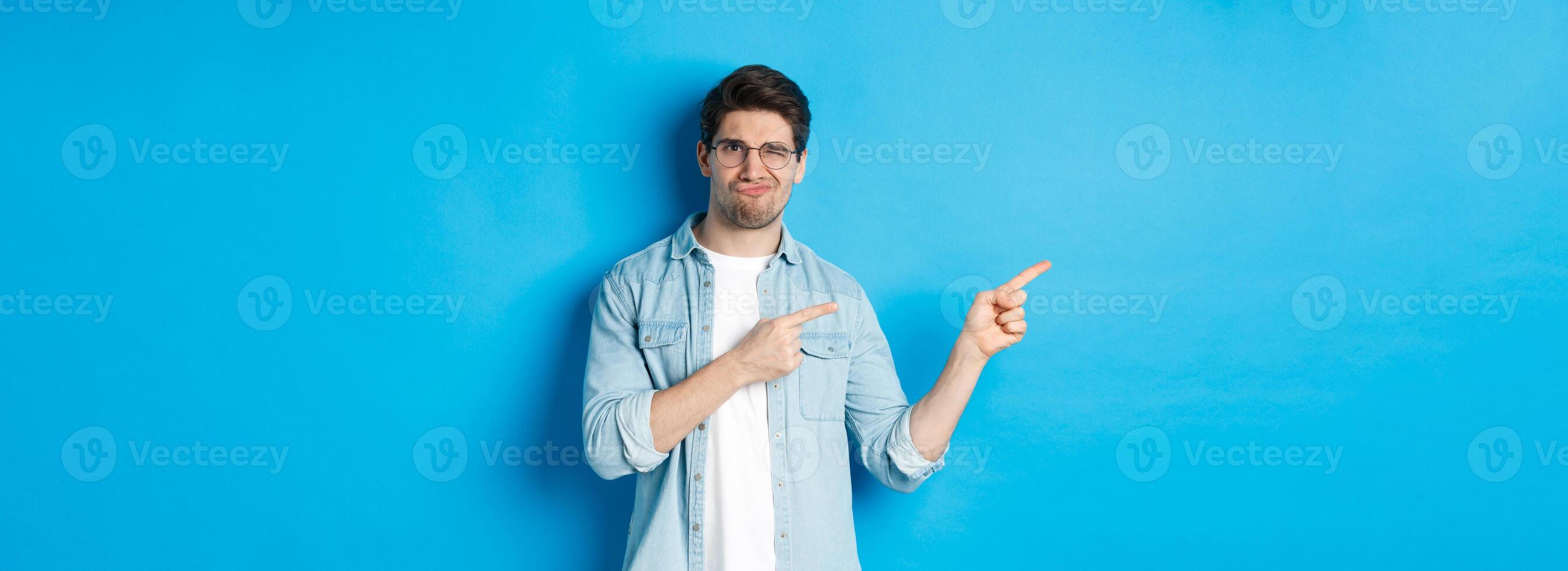 Displeased and skeptical male model in glasses, pointing fingers left at something bad, showing awful advertisement, standing over blue background photo