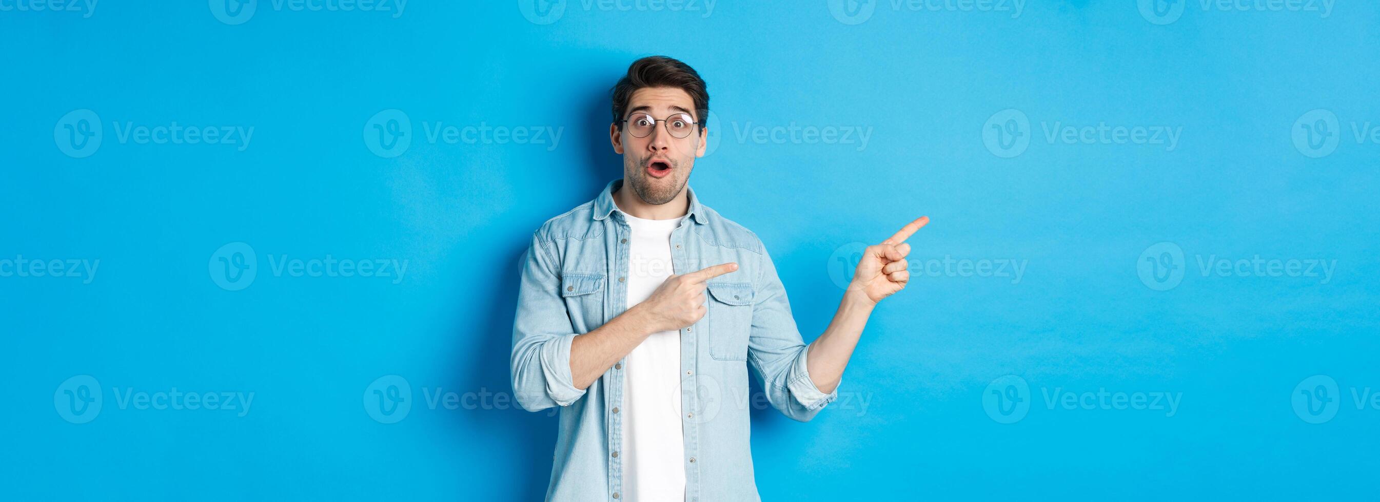 Surprised man in glasses pointing fingers left at copy space, asking question about product, looking intrigued, standing over blue background photo