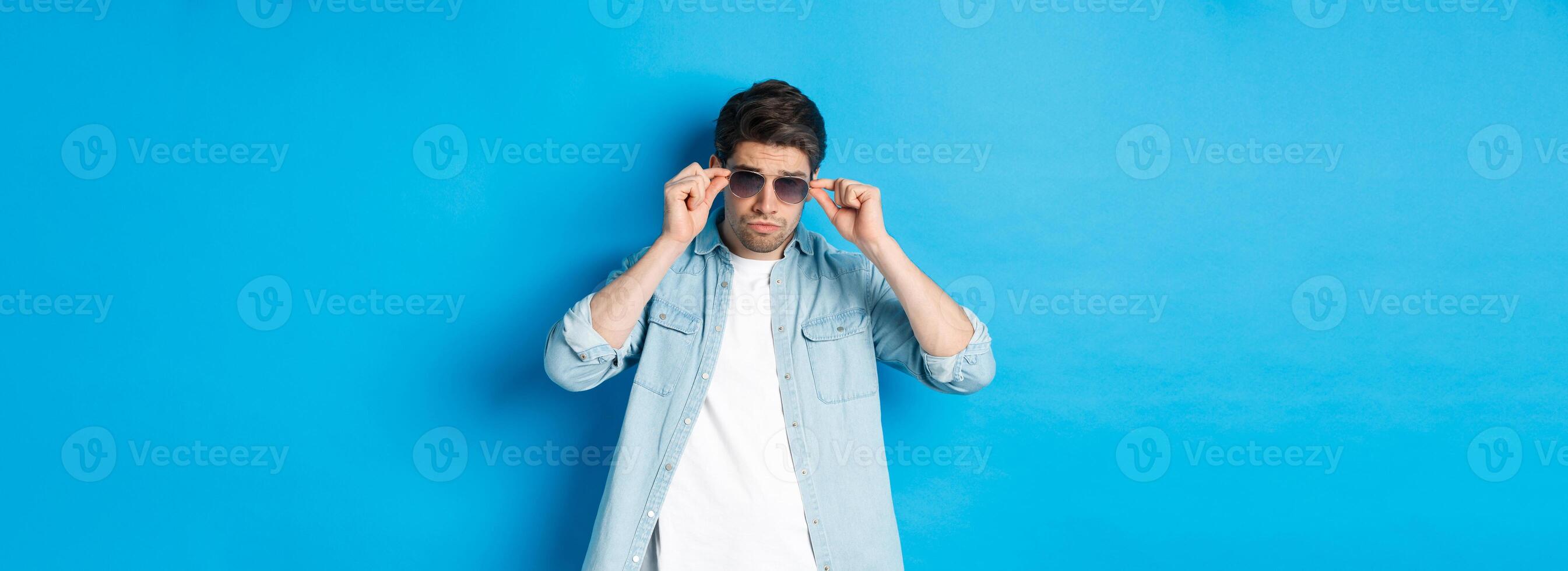Confident macho man put on sunglasses, looking cool and sassy, standing over blue background photo