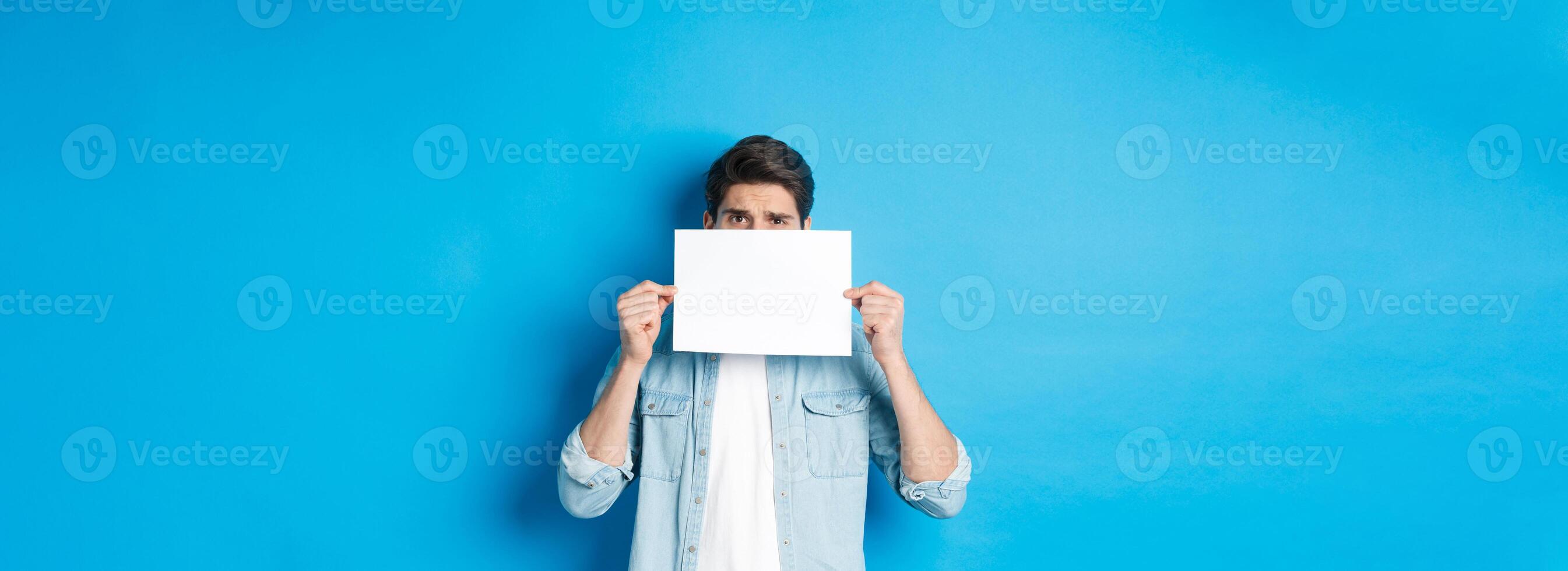 Shy and nervous guy hiding face behind blank piece of paper, peeking with worried look, showing your logo, standing over blue background photo