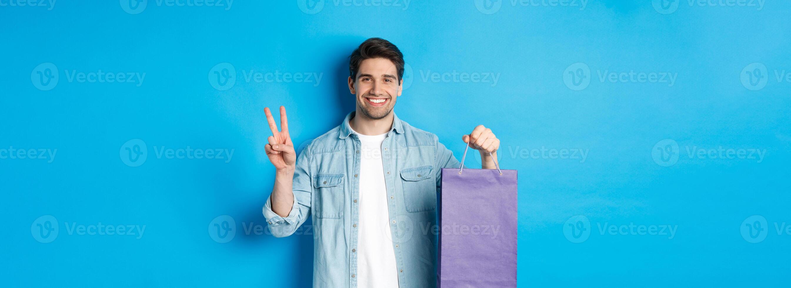 Concept of shopping, holidays and lifestyle. Happy bearded guy holding paper bag from store and showing peace sign, standing over blue background photo