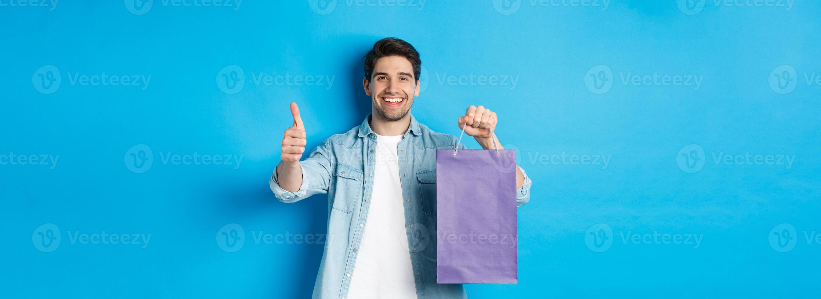 Concept of shopping, holidays and lifestyle. Satisfied smiling man holding paper bag, showing thumb-up and recommending store, standing over blue background photo