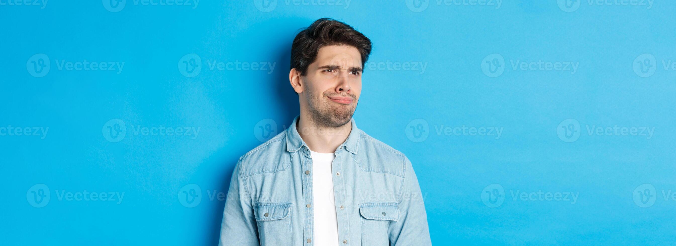 Close-up of upset and disappointed man grimacing, looking dissatisfied left, cringe from bad promo banner, standing over blue background photo