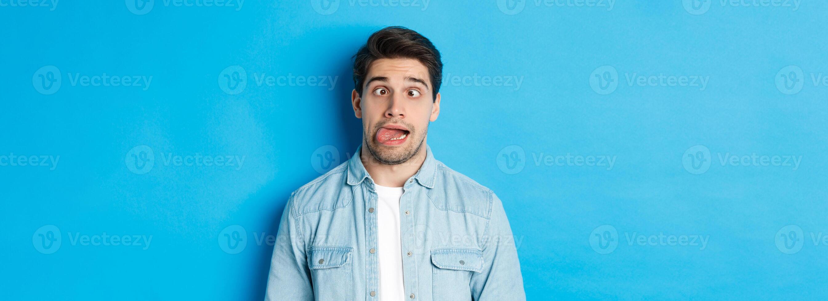 Close-up of young man making funny expressions, showing tongue and looking at camera, standing over blue background photo
