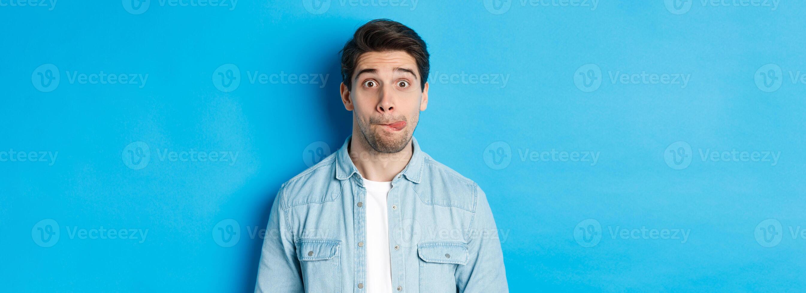 Close-up of young man making funny expressions, showing tongue and looking at camera, standing over blue background photo