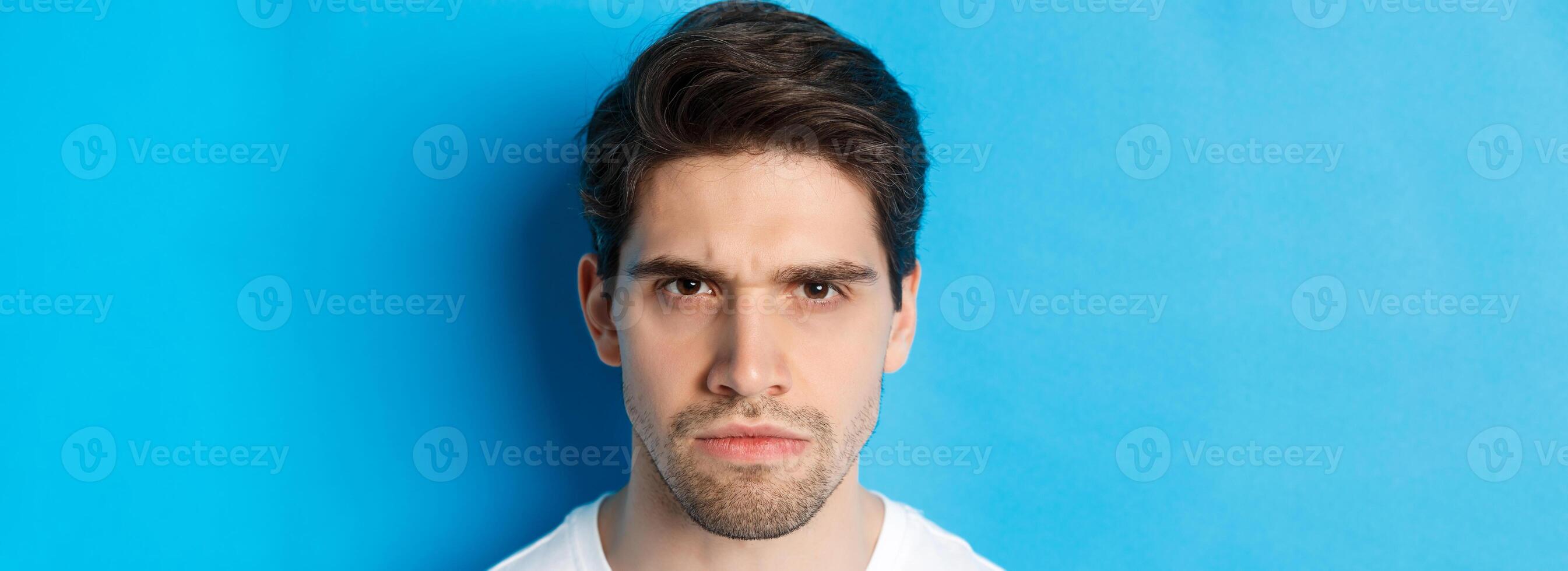 Headshot of angry man frowning, looking disappointed and bothered, standing over blue background photo