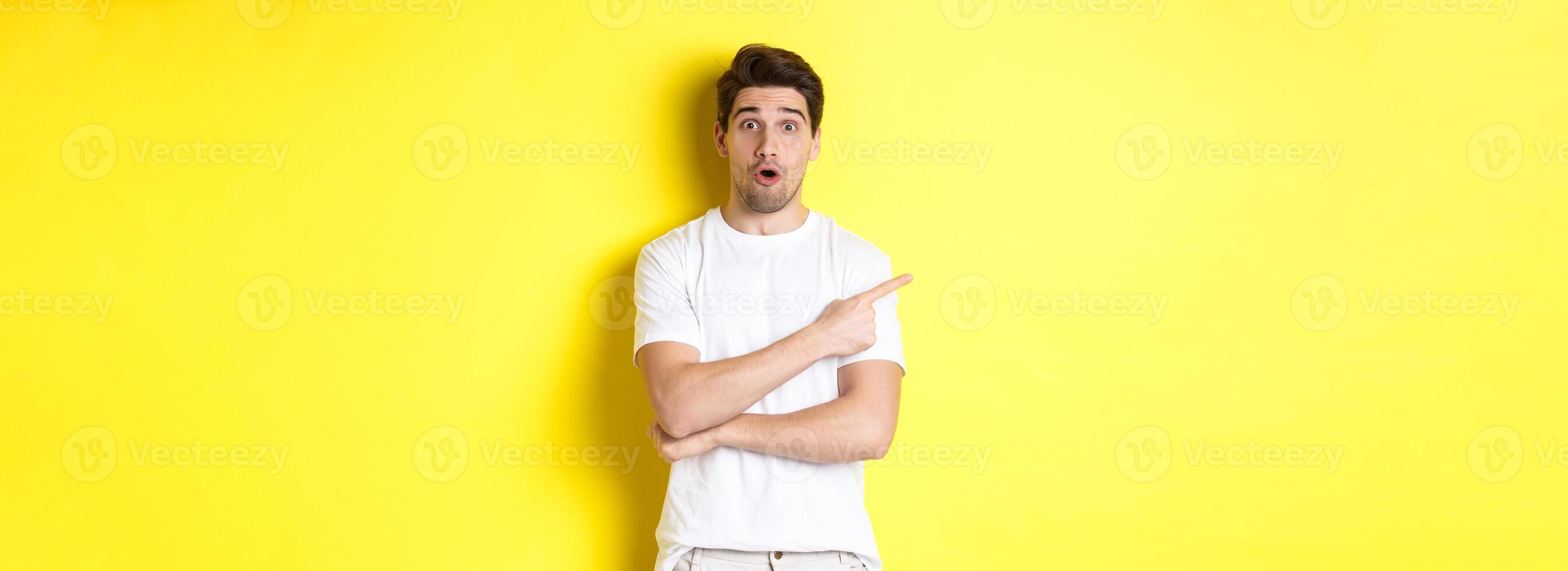 Cuious guy pointing finger left, asking question about promo offer, showing banner, standing amazed against yellow background photo