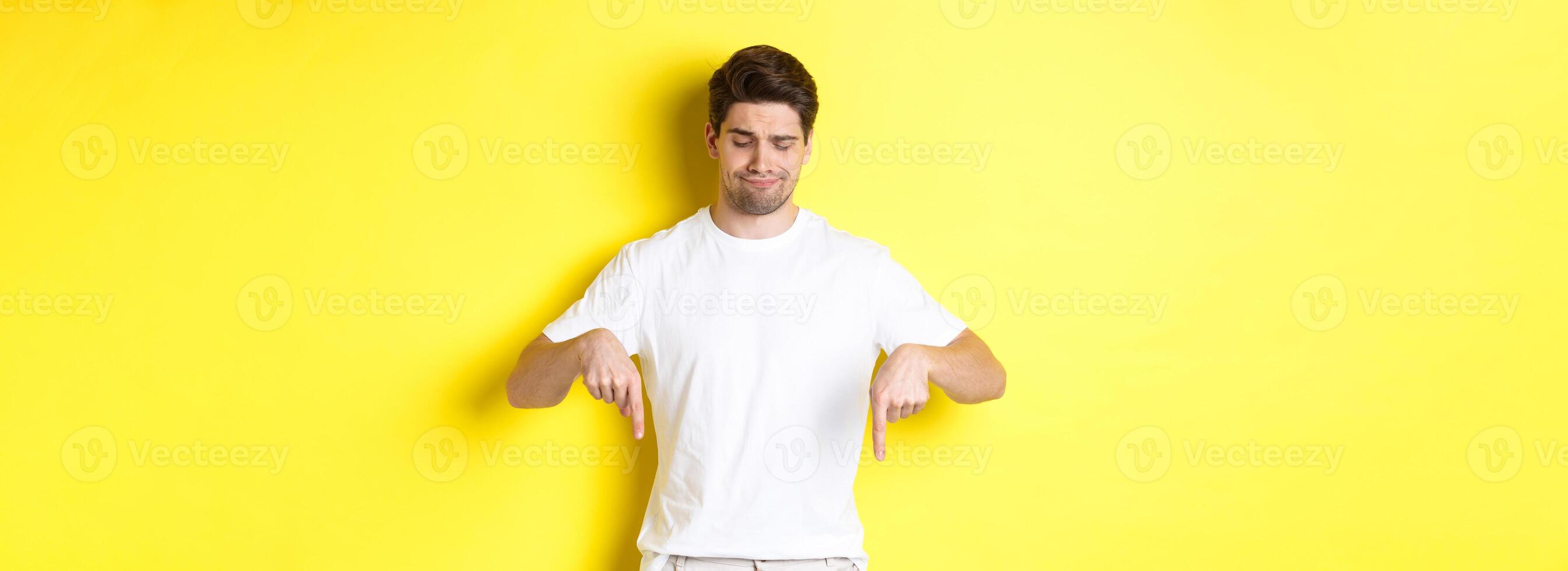 Skeptical young man in white t-shirt, pointing and looking down upset, disapprove and dislike product, standing over yellow background photo