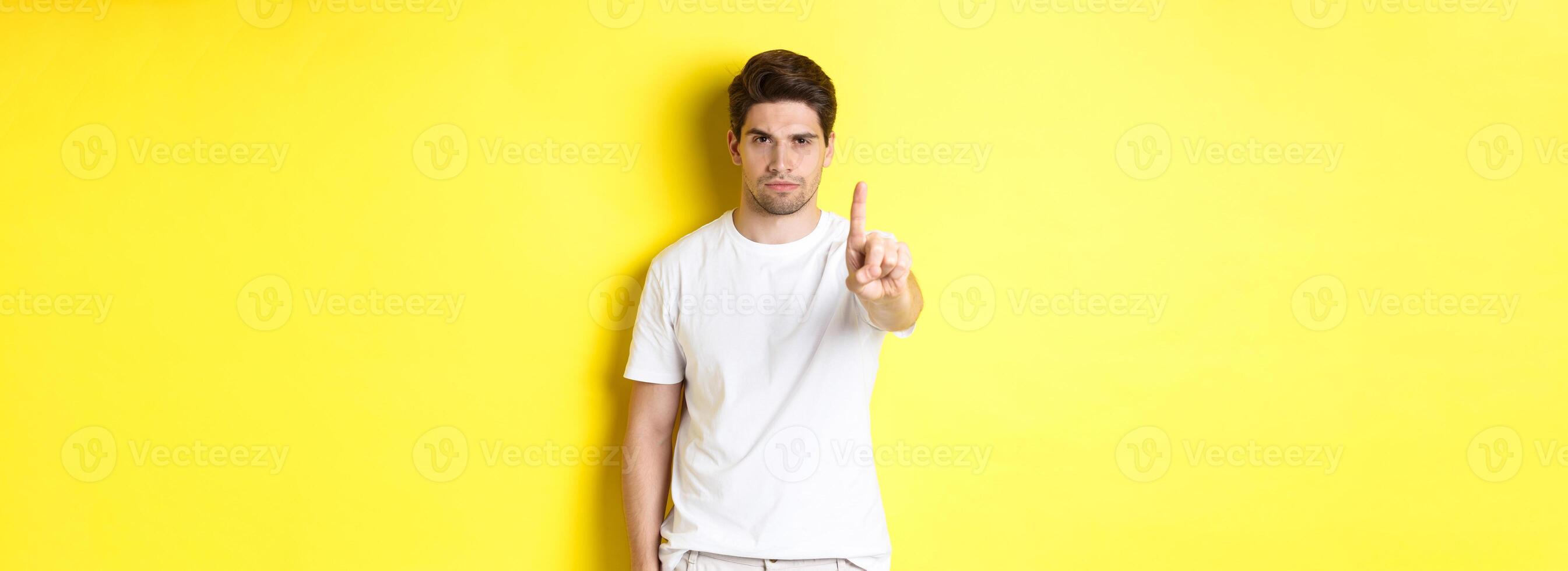 Confident man saying no, decline and prohibit something, showing one finger and frowning, standing over yellow background photo