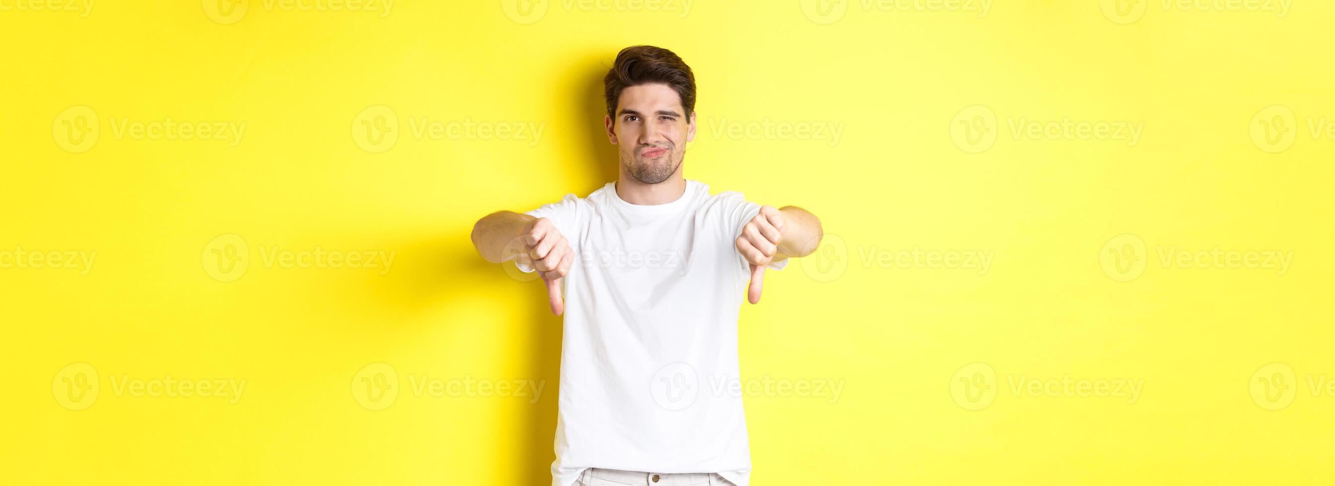 Skeptical guy showing thumbs down and smirking, dislike and disagree, standing over yellow background photo