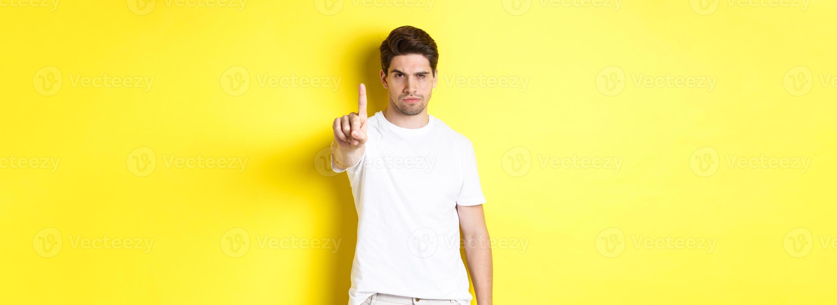 Serious man frowning displeased, shaking one finger to prohibit something, warn you, standing over yellow background photo