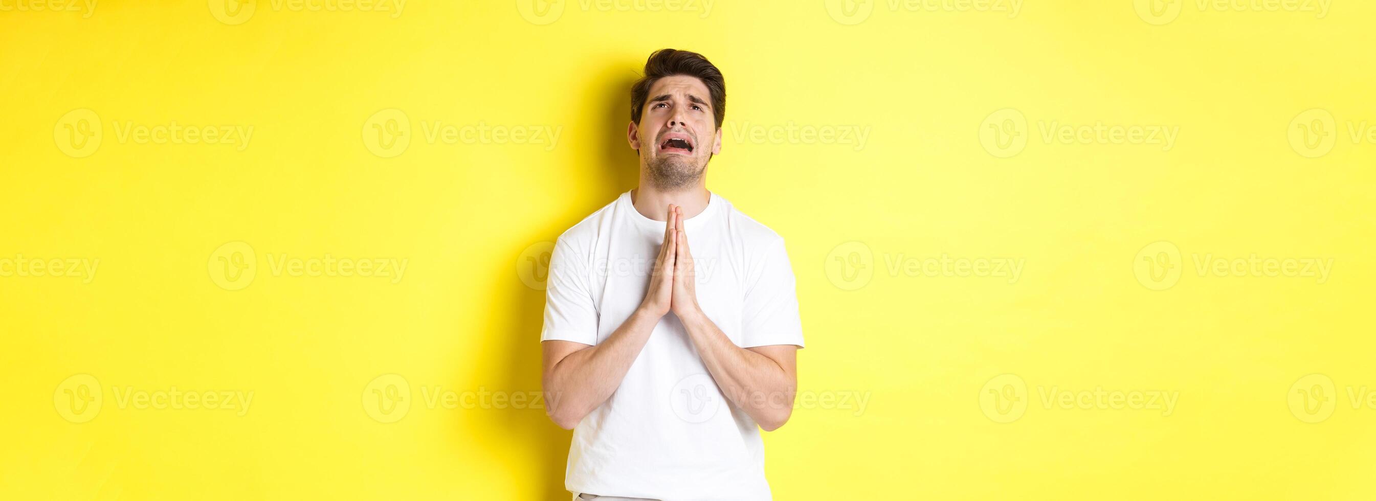 Desperate man pleading God, holding hands in pray and looking up distressed, standing over yellow background photo